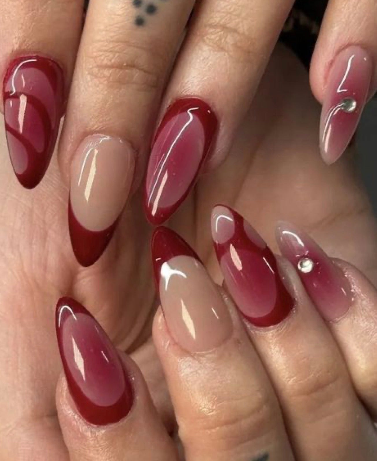 Close-up of manicured fingernails with almond-shaped tips, featuring a glossy dark red ombre gradient transitioning into a sheer nude-pink base, with one nail decorated by a small rhinestone.