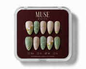 MUSE nails press-on set in a clear plastic clamshell case with a dark burgundy insert. The set features almond-shaped nails in beige and sage green with gold leaf and floral details, with size options XS, S, M, and L indicated.