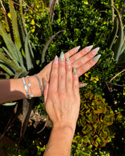 A pair of hands with long, sculpted stiletto nails painted in a pale sage green glitter and clear gradient design