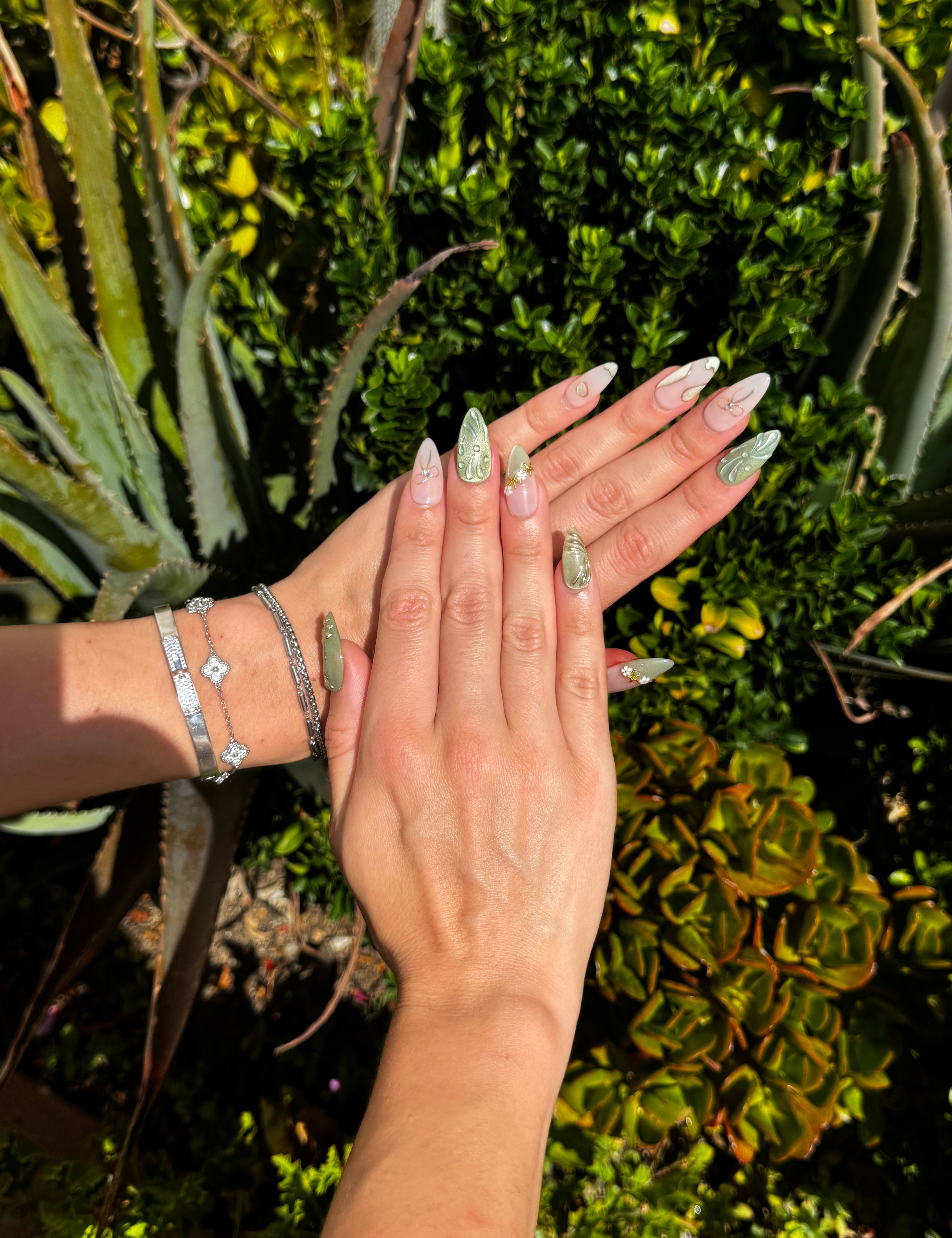 A pair of hands with long, sculpted stiletto nails painted in a pale sage green glitter and clear gradient design