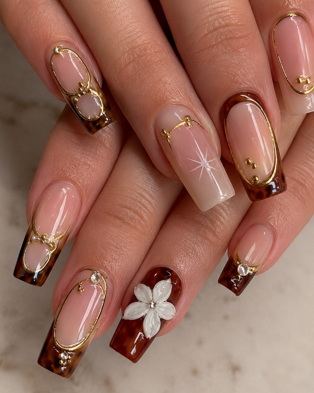 Close-up of manicured fingernails with a glossy nude-pink base, dark brown French tips, gold metallic detailing, and one white flower accent on a light marble surface.