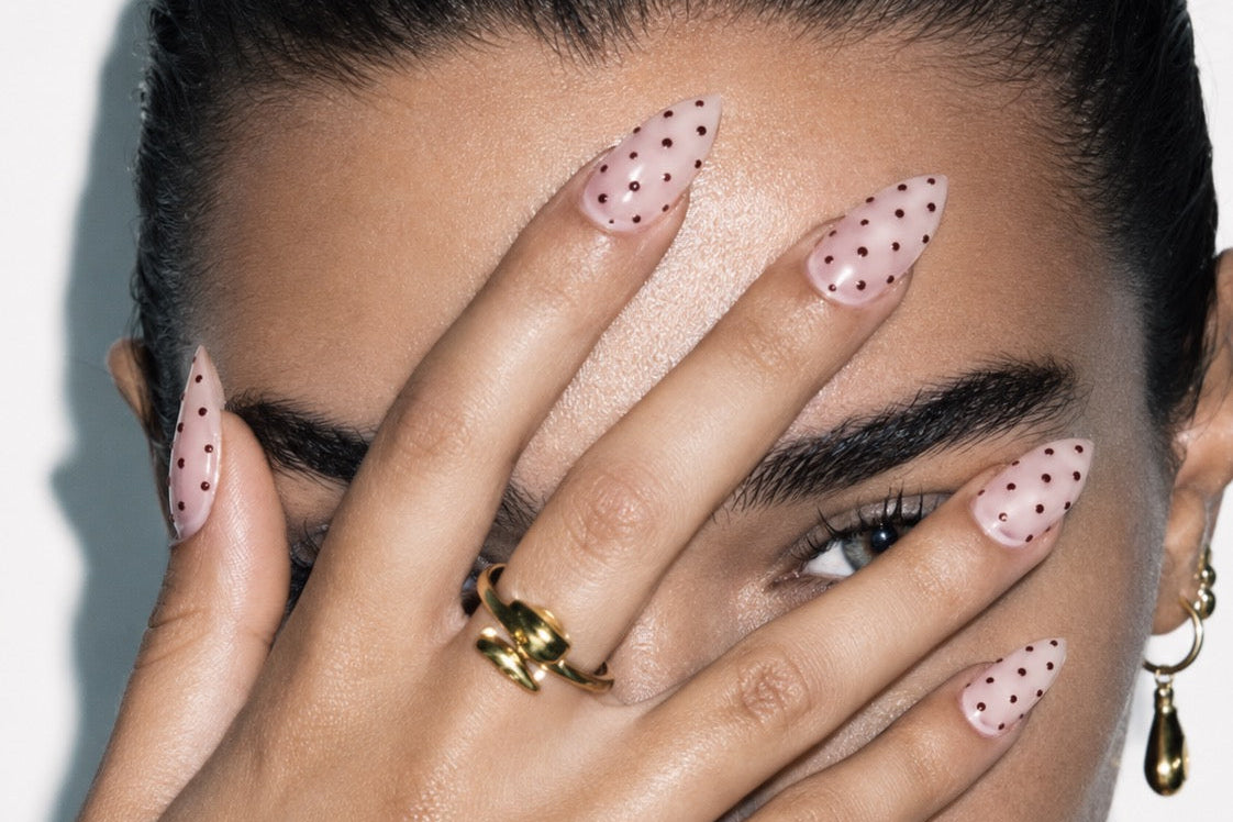 A close-up of a model's hand with long, almond-shaped pink nails decorated with small black polka dots, covering part of their face. The model has dark hair pulled back and is wearing a gold ring and hoop earring against a plain white background.