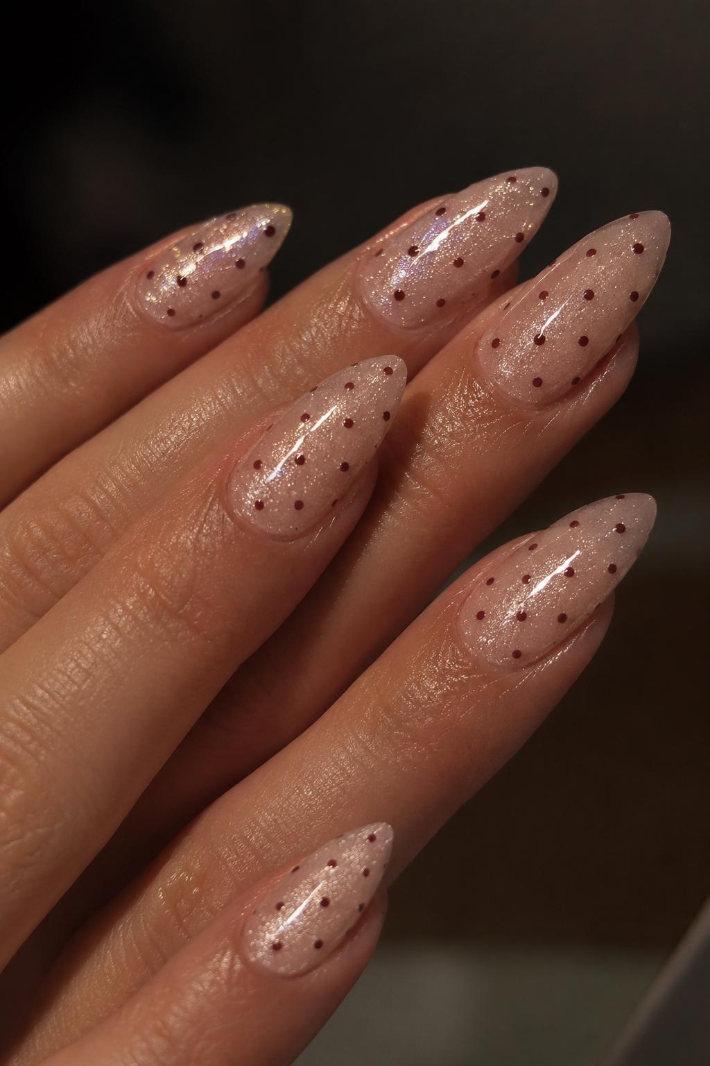 Close-up of a hand with long, almond-shaped nails painted in a sheer nude pink with small dark red polka dots, against a dark blurred background.