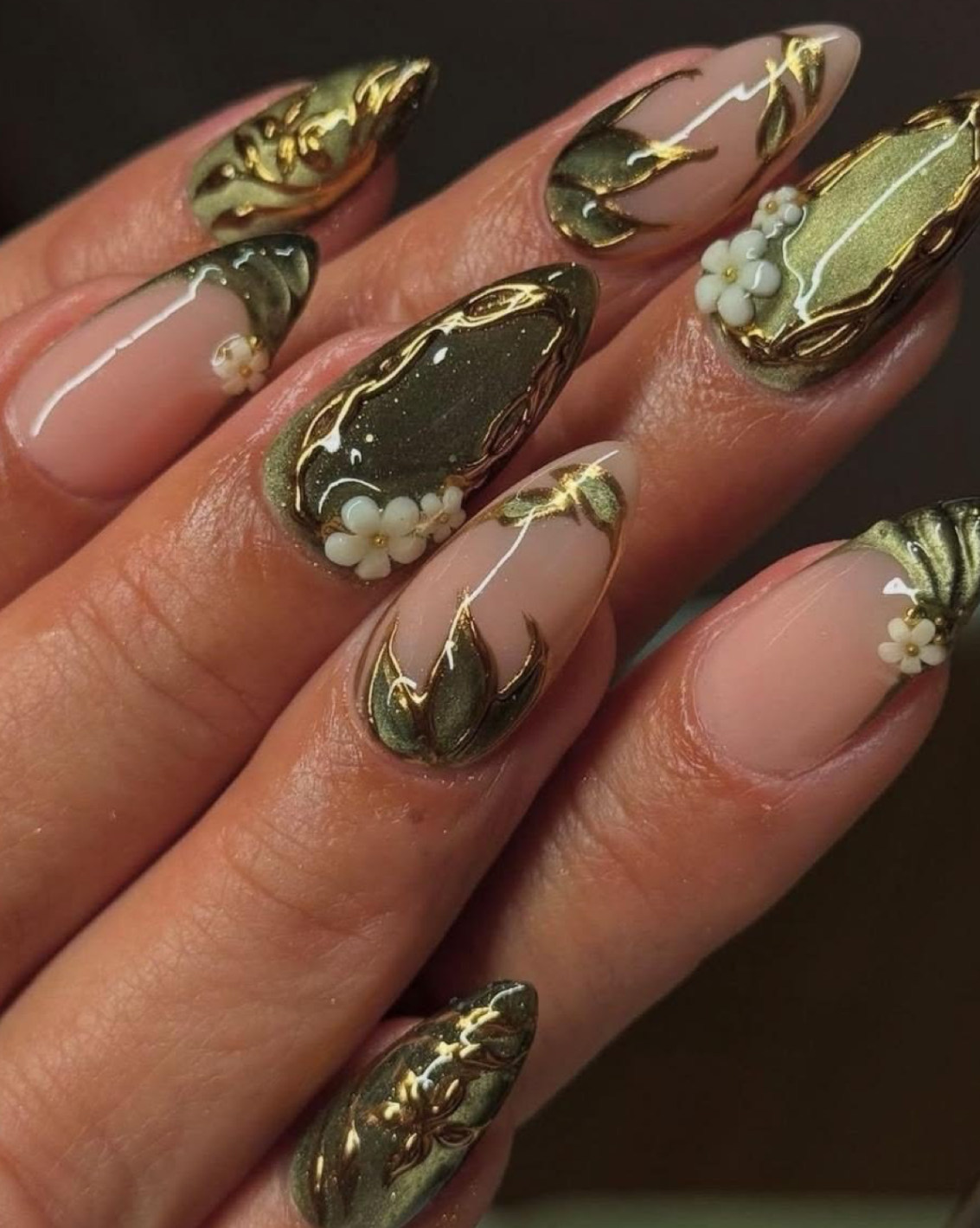 A close-up of manicured fingernails with a glossy nude-pink base, metallic olive green and gold leaf-like patterns, and small white pearl accents, shown on a hand against a dark background.