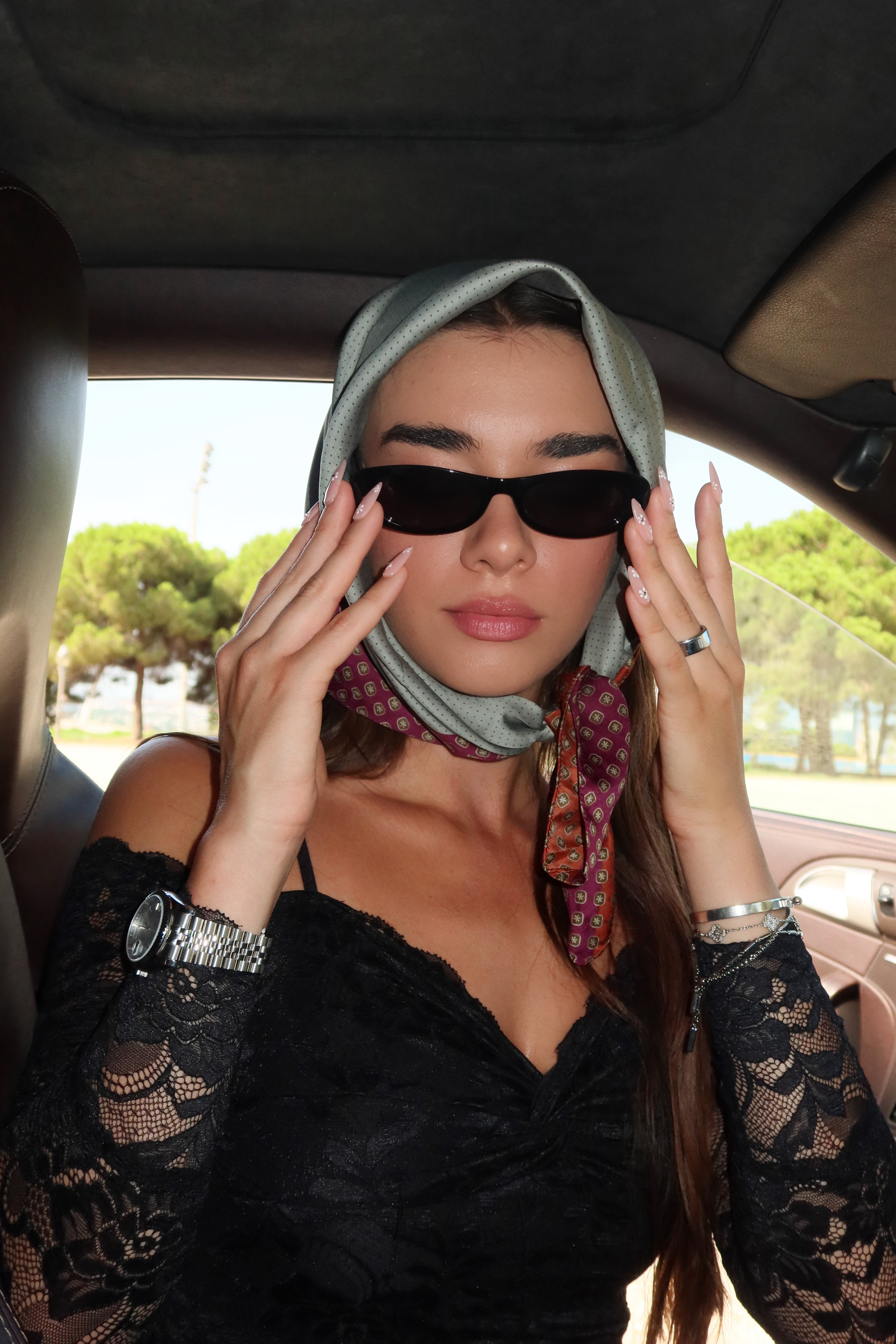A woman wearing a black lace top, black sunglasses, and a gray patterned headscarf sits inside a car, holding the sunglasses in front of her eyes with press on nails from muse set daisy