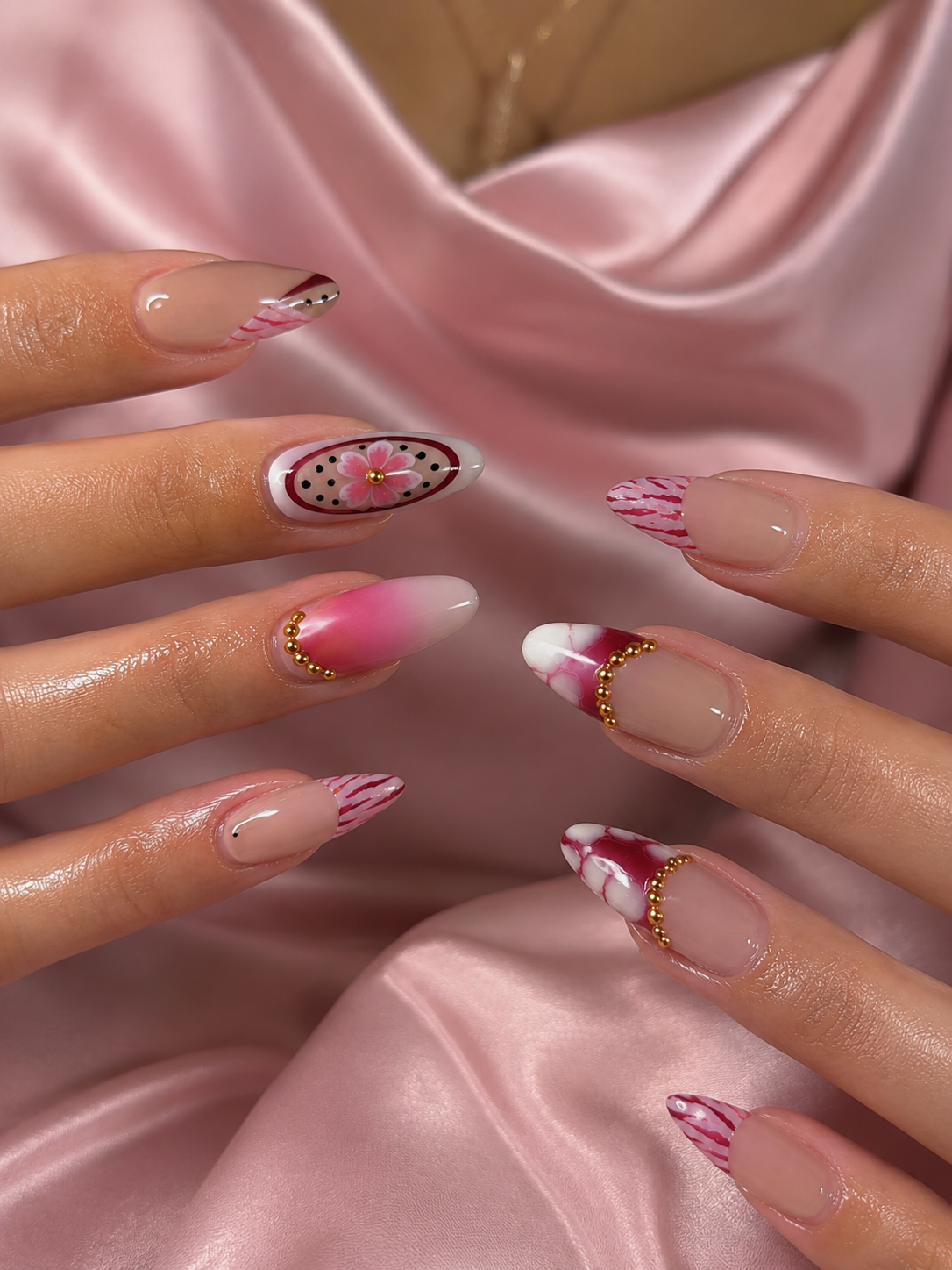 Close-up of manicured fingernails with pink and nude designs, shown against a soft pink satin fabric background.