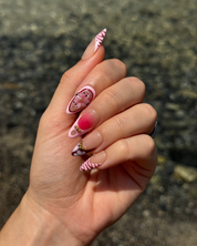 A close-up of a hand showcasing a pink and white almond nail art set with various designs including stripes, a pink gradient, pink flowers, and small gold glitter accents, set against a blurred outdoor background.