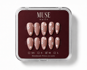 A set of 10 press-on nails by MUSE nails, displayed in a clear plastic clamshell case with a dark burgundy insert. The nails are a warm nude pink with white floral patterns, and the size 'M' is checked on the size guide at the bottom.